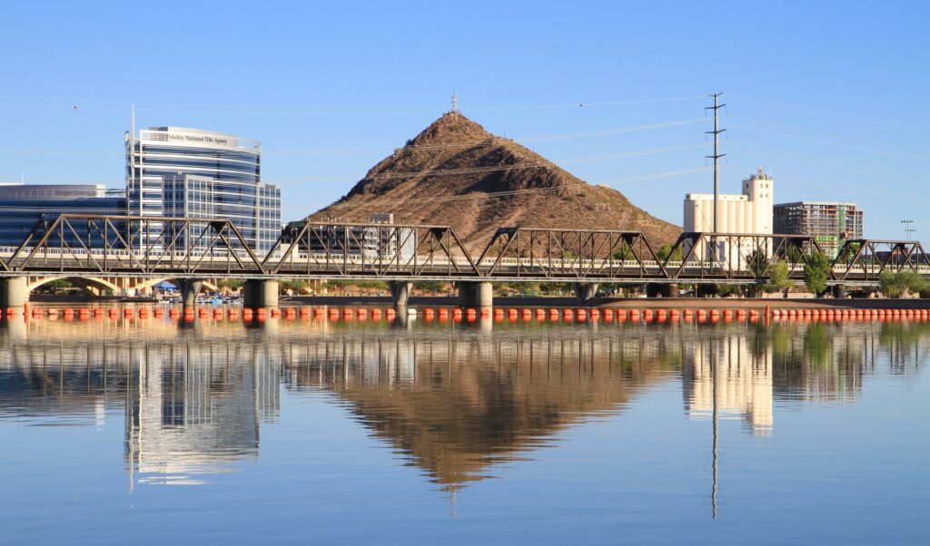 View of a waterfront corridor featuring a steel truss bridge crossing a calm lake with reflections of nearby buildings and a distinctive desert butte.
