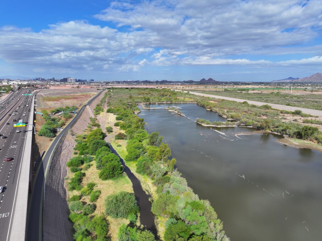 Aerial view of a river corridor featuring a winding waterway bordered by dense desert vegetation and a paved multi-use trail. A major freeway runs parallel to the river on the left, while open desert, distant mountains, and the urban skyline extend into the background under a partly cloudy sky.