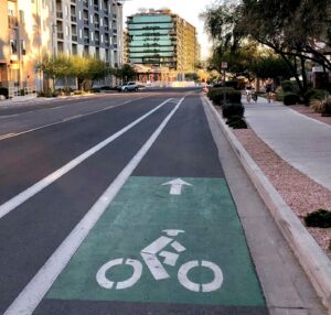 Multi-lane street in the middle of busy city. On the right side a bike line is positioned between the sidewalk and the lanes for cars. Between the bike path and cars, a buffer lane is present. 