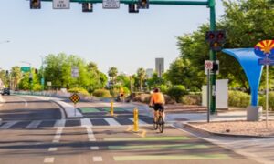 A multi-lane street intersection surrounded by green trees on either side. A biker is riding on the right side of the double lane bike path.