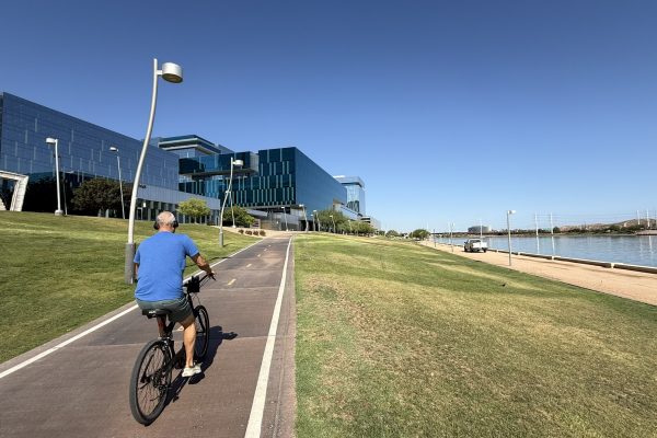 A cyclist rides along a paved waterfront shared-use path bordered by open green space and modern office buildings. The path is lined with regularly spaced lighting.