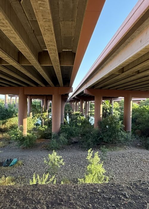 A riverside trail lined with decorative lighting runs alongside an elevated freeway structure and a fenced waterway.