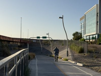 A cyclist rides along a paved multi-use path within an urban corridor bordered by freeway infrastructure and modern office buildings. Decorative lighting, fencing, and landscaped desert vegetation line the pathway, which provides a separated route for walking and biking through a developed area.