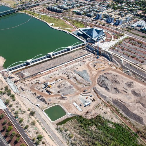 aerial view of an urban waterfront area featuring a lake, bridges, trails, and surrounding mixed-use development. Along the shoreline, A pedestrian bridge crosses the water. Residential neighborhoods, roadways, and landscaped open spaces surround the waterfront.