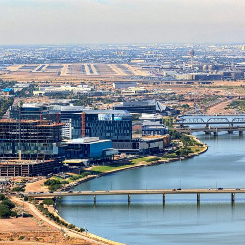Aerial view of a vibrant waterfront district featuring modern office buildings, mixed-use development, bridges, and recreational amenities surrounding a man-made lake.