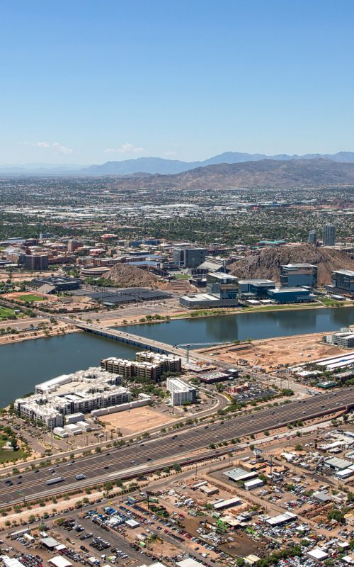 Aerial view of an urban waterfront corridor featuring a series of lakes, bridges, mixed-use developments, and transportation infrastructure set within a desert metropolitan landscape.